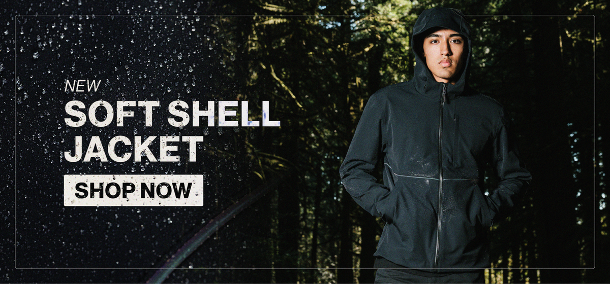 New Soft Shell Jacket. A model stands in a dark, misty forest wearing a black hooded soft shell jacket. The hood is up, the jacket is zipped, his hands are in the pockets, and water droplets are beaded on the fabric.